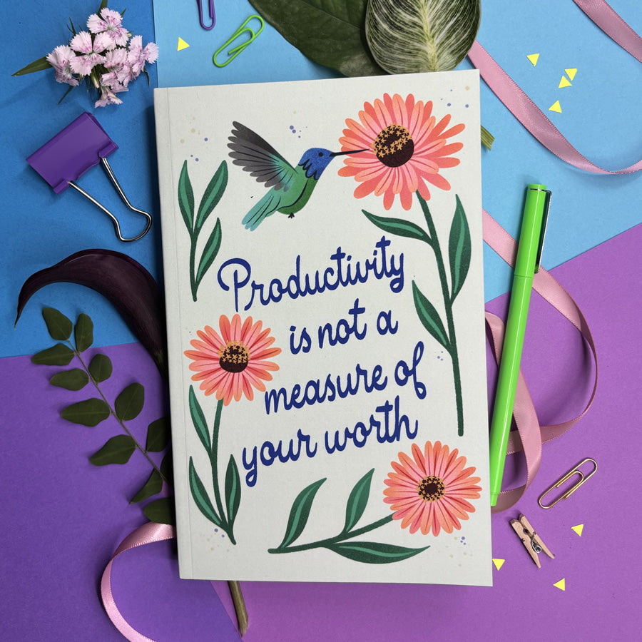 Notebook with lettering "productivity is not a measure of your worth" with a hummingbird and flowers surrounded by stationery items on a colorful background