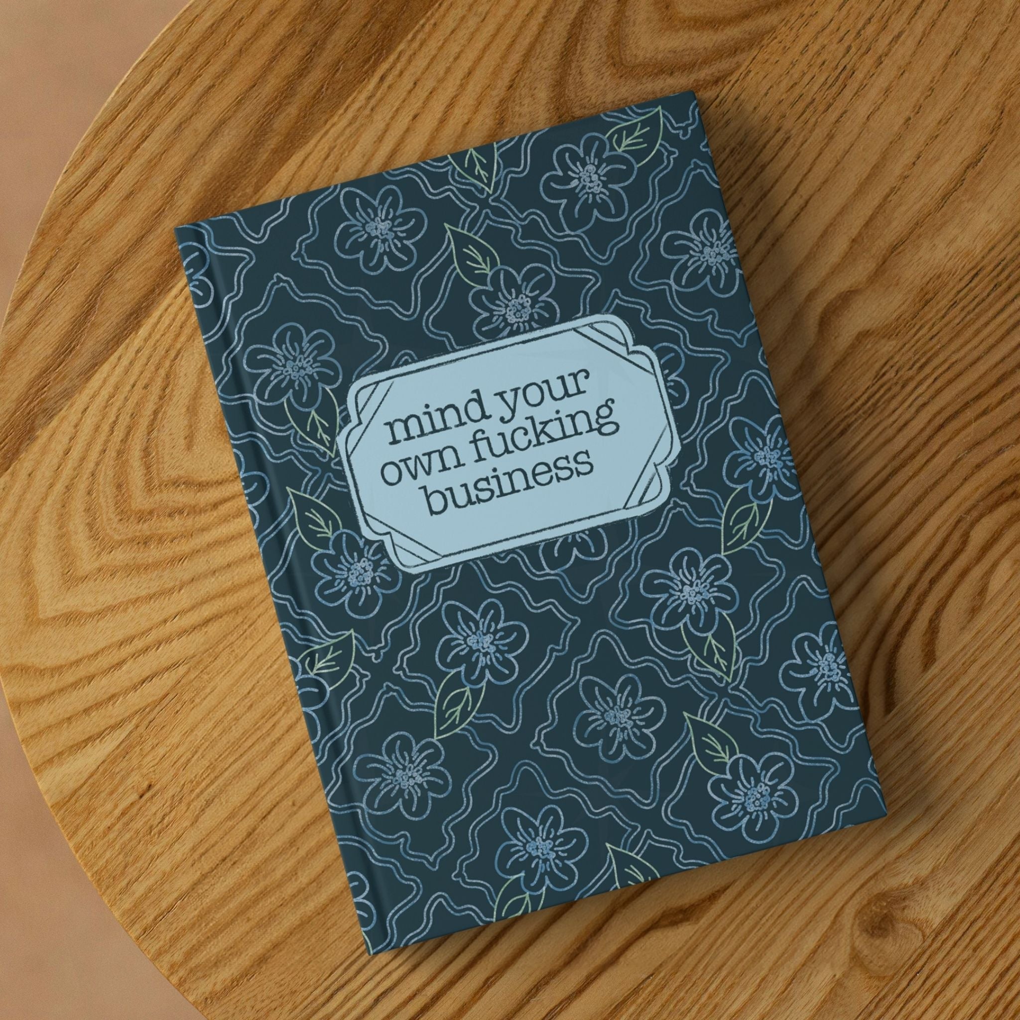 Profanity floral hardcover journal lying on round wooden surface showing front cover with teal botanical pattern