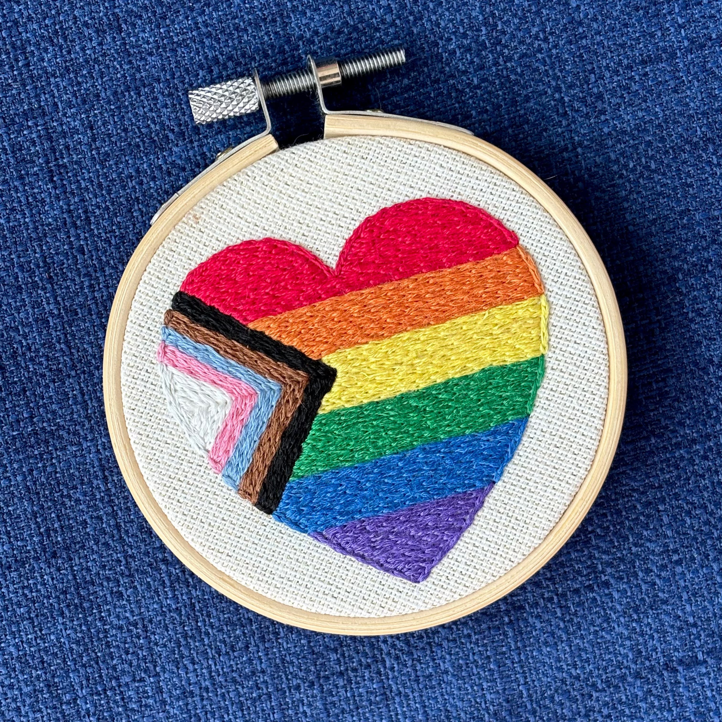 Heart-shaped progress pride flag hand embroidered onto cream colored linen. Displayed in a light wooden embroidery hoop as wall art or a christmas ornament.