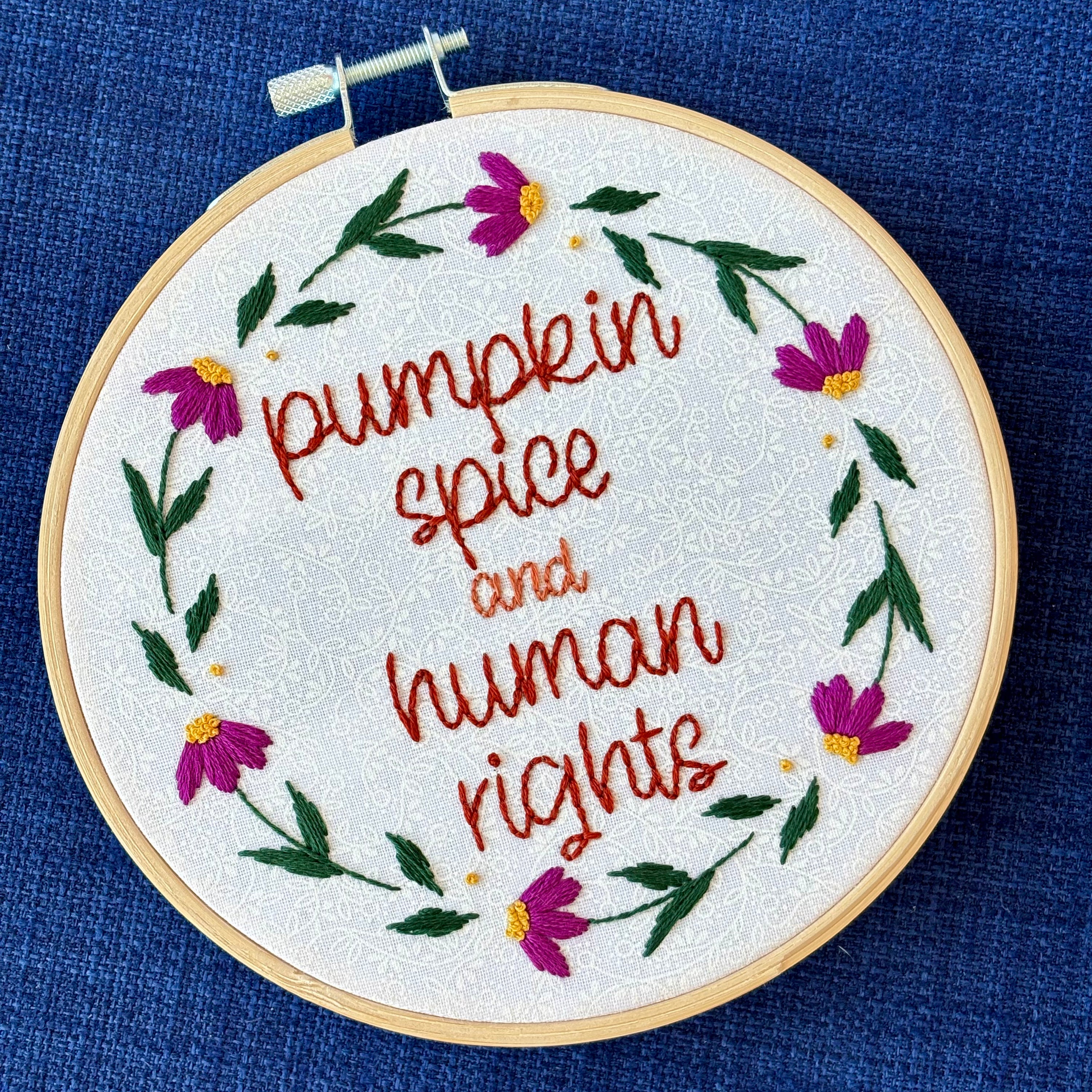 "Pumpkin spice and human rights". Handwriting font text hand embroidered in two shades of medium orange. Surrounded by a floral wreath of bright pink flowers with yellow centers and green leaves and stems. Framed in light wood embroidery hoop and displayed as wall art.