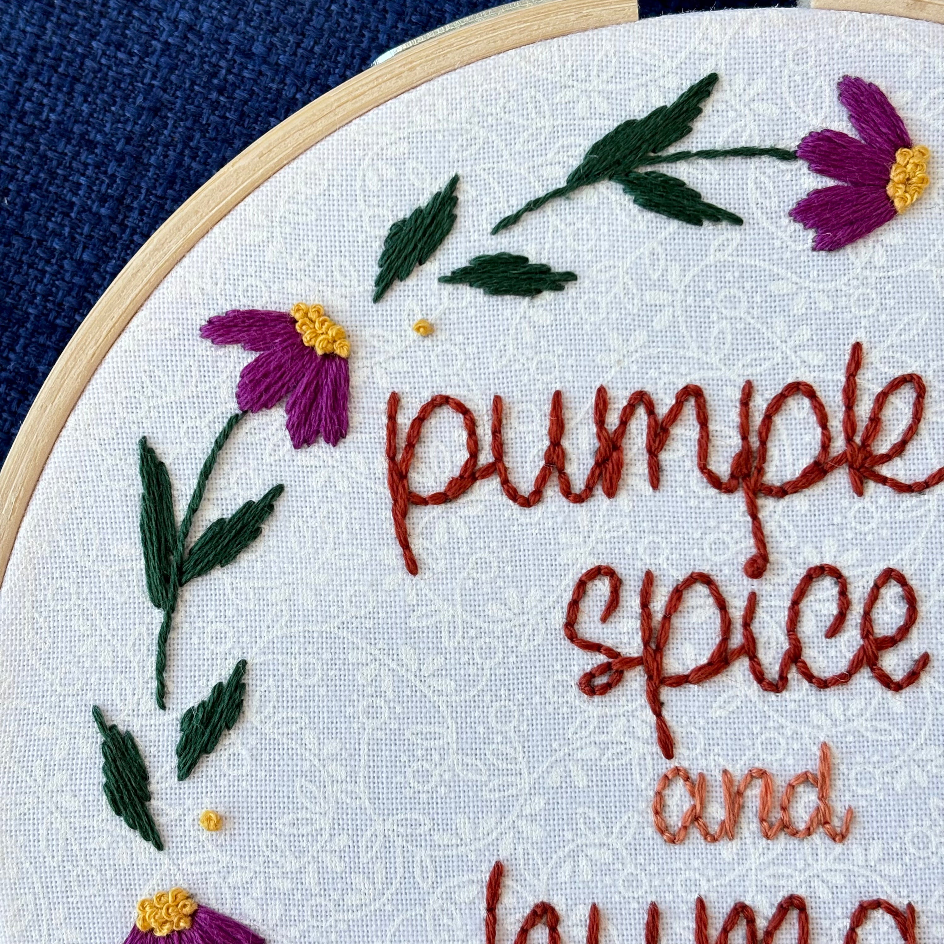 "Pumpkin spice and human rights". Handwriting font text hand embroidered in two shades of medium orange. Surrounded by a floral wreath of bright pink flowers with yellow centers and green leaves and stems. Framed in light wood embroidery hoop and displayed as wall art.