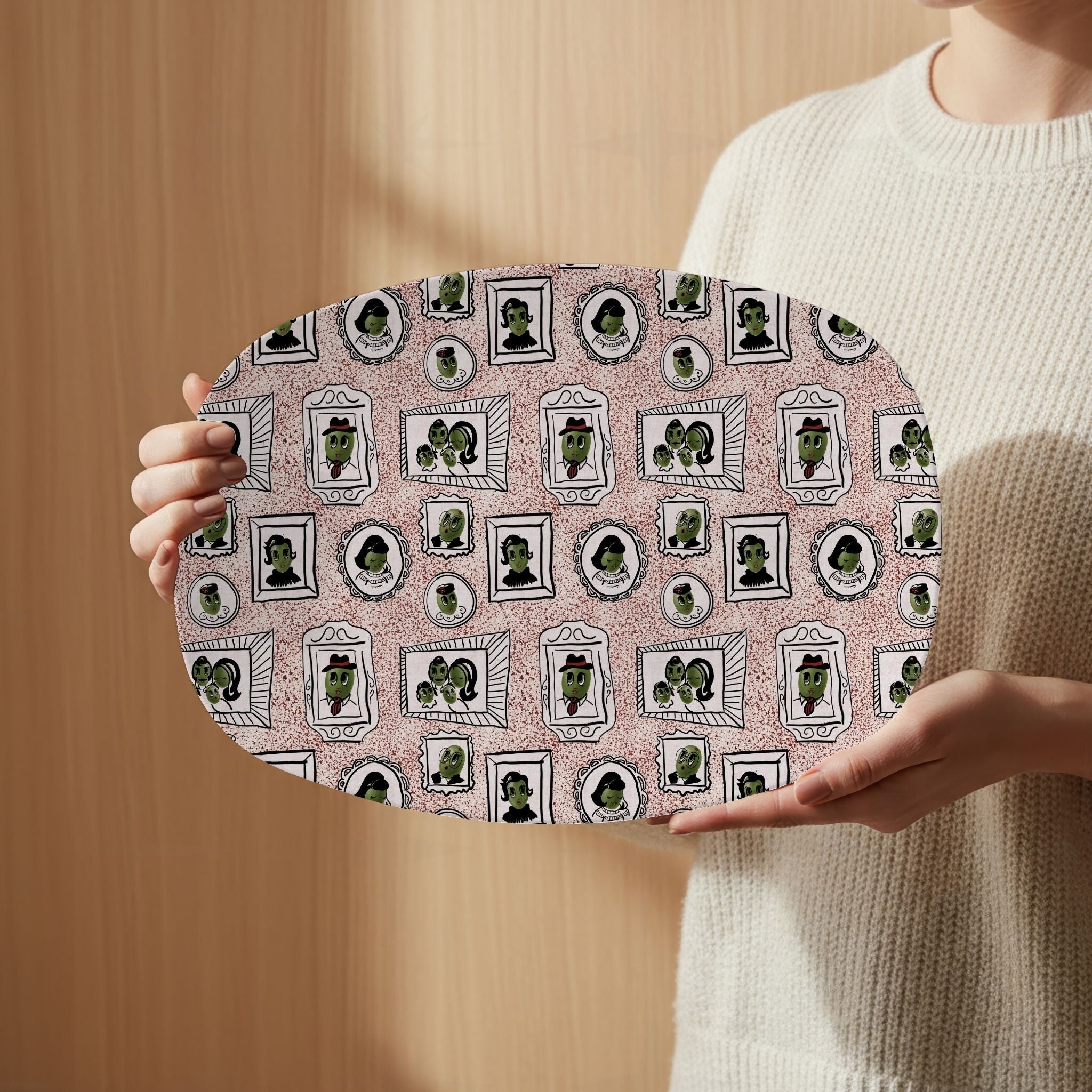 Person in cream sweater holding retro olive portrait serving platter in red speckle colorway face-forward, full mid-century gallery wall framed olive pattern visible against warm wood background