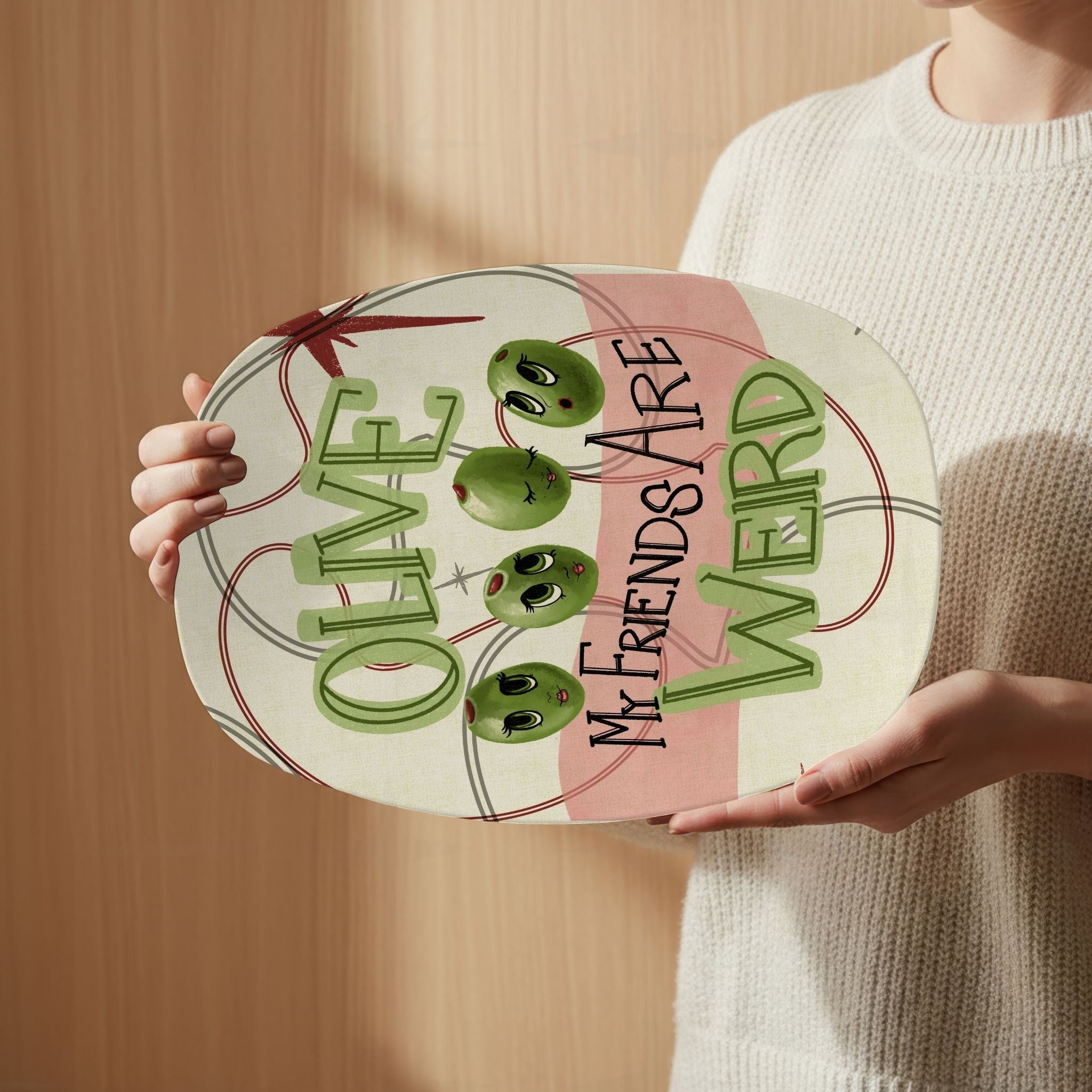 Person in cream sweater holding retro olive pun serving platter showing full illustrated mid-century modern design with four olive characters and "Olive my friends are weird" lettering, warm wood background
