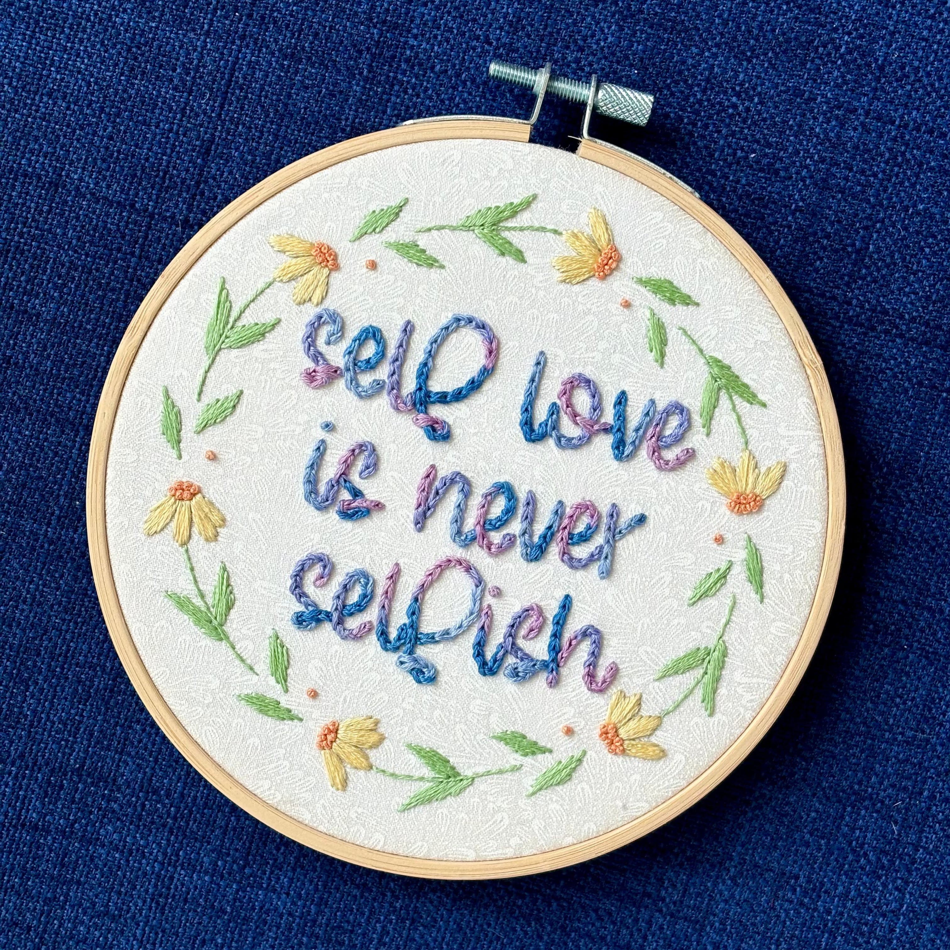 "self love is never selfish." Quote hand embroidered in color changing thread of pink, lavender, light blue, and royal blue. Surrounding the quote is a floral wreath of yellow and orange daisies. Sewn on white-on-white cotton. Finished wall art is framed in a light wood embroidery hoop.