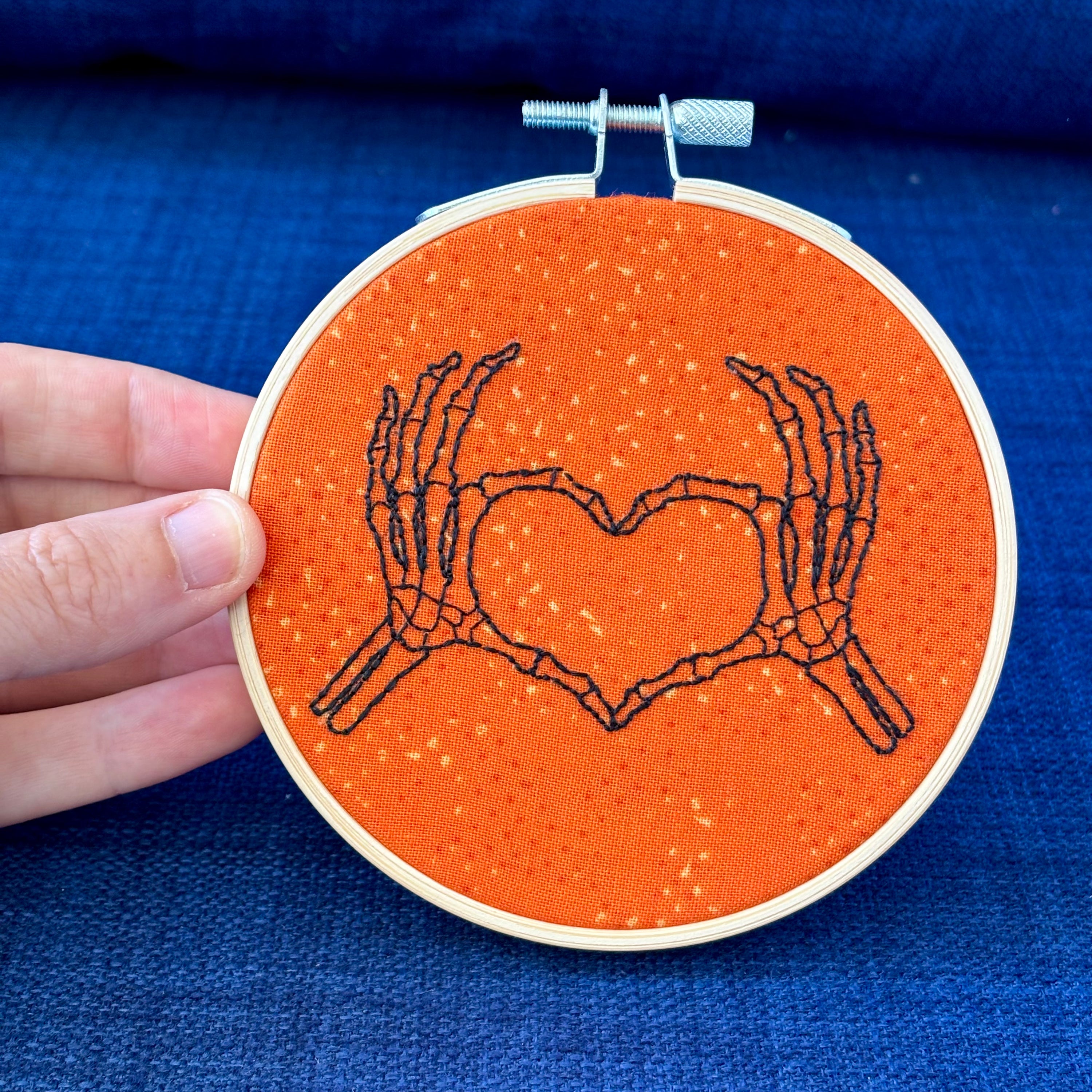Hand embroidered pair of skeleton hands making a heart by touching thumbs and forefingers, while remaining fingers fan out. Bones are outlined with fine black embroidery. Stitched on bright orange cotton and displayed in light wooden hoop as wall art or Christmas ornament