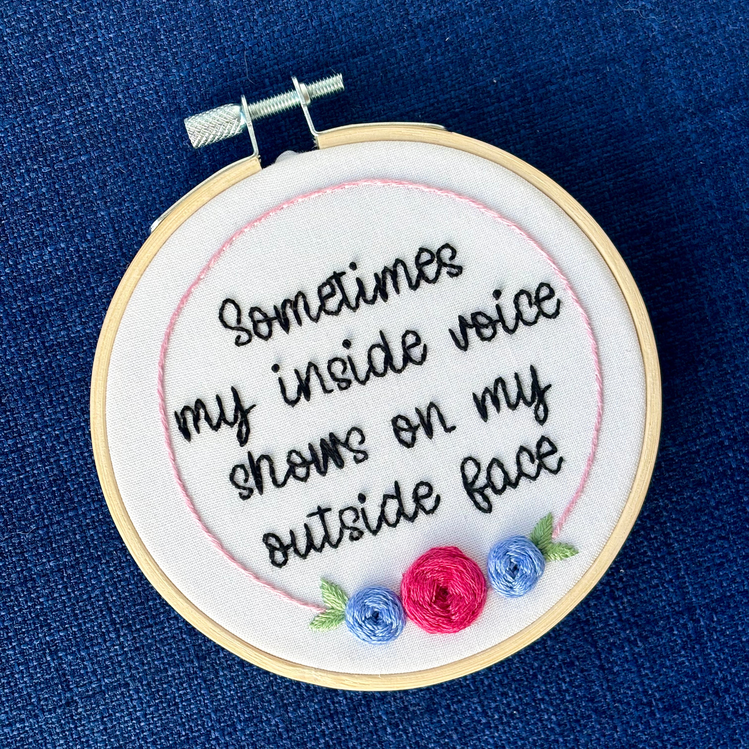 Completed embroidery hoop reads, "Sometimes my inside voice shows on my outside face". Quote in handwriting font and hand embroidered in black. Words are centered in a floral frame, which is light pink and embellished with cheerful fuchsia and periwinkle roses and light green leaves sewn with embroidery thread. Fabric is white cotton. Framed in light wood embroidery hoop and displayed as wall art or Christmas ornament.