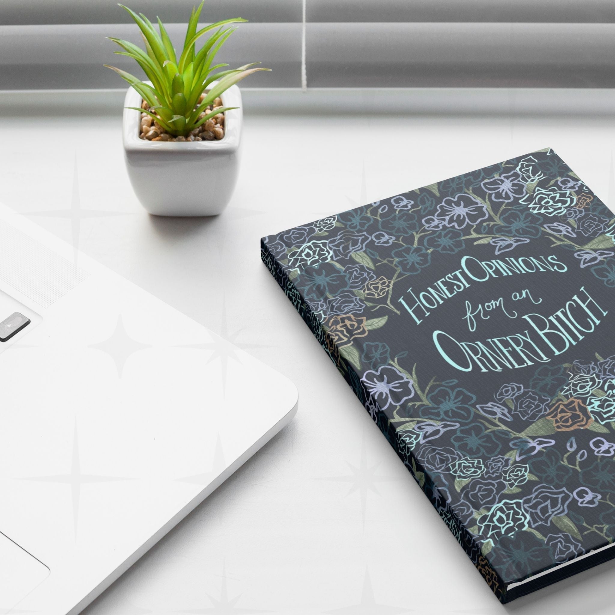 Sweary dark floral hardcover journal on white desk beside laptop and small potted succulent, lifestyle setting