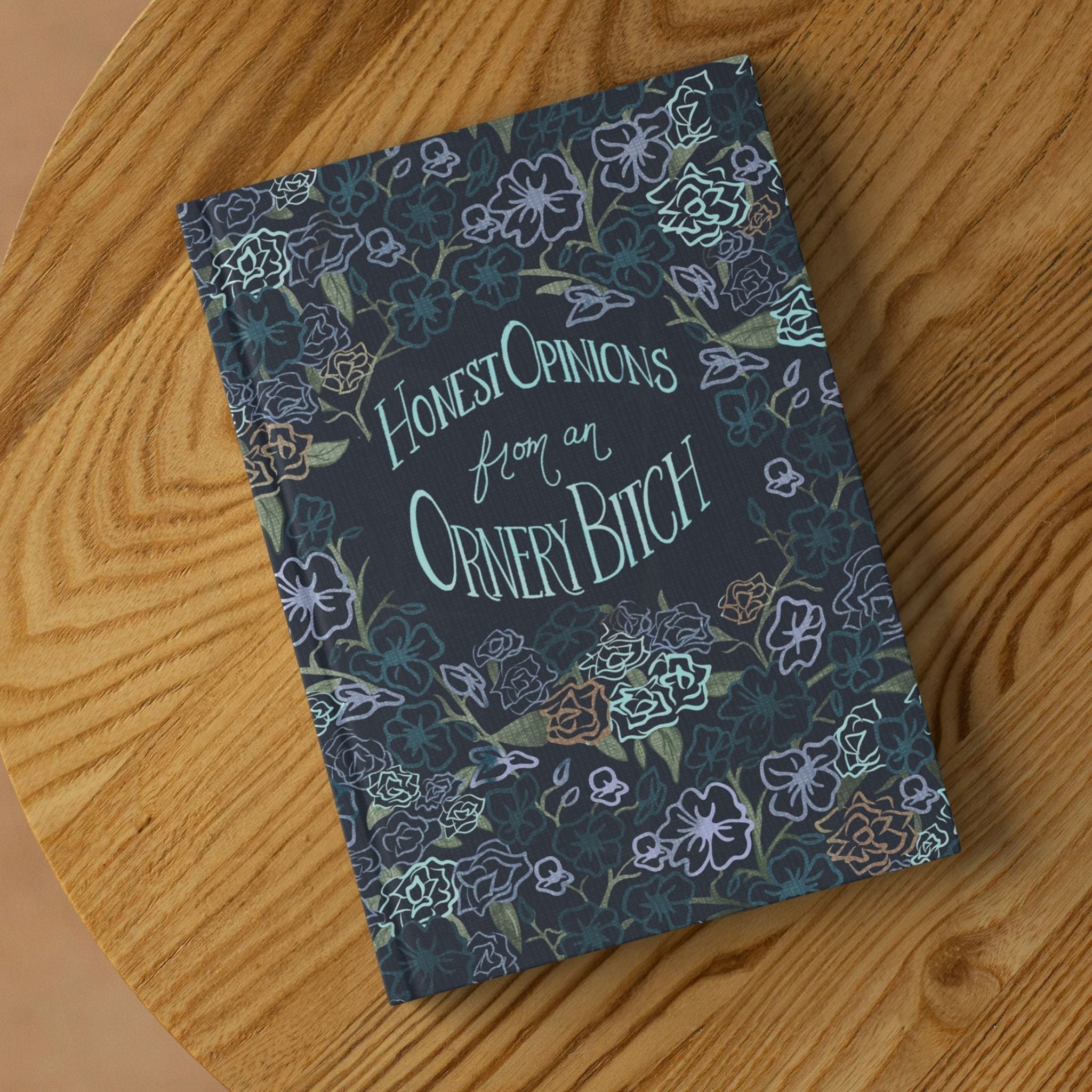 Sweary dark floral hardcover journal lying on round wooden surface showing front cover with hand-lettered Honest Opinions from an Ornery Bitch text and gothic botanical pattern