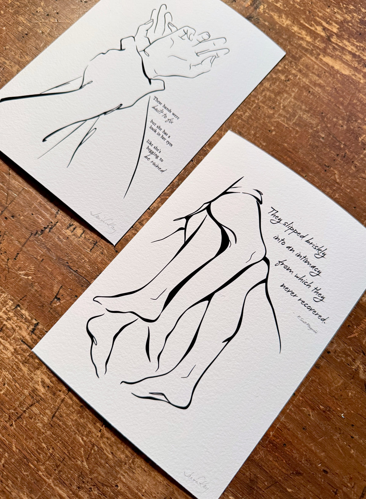 Fine art prints of "I Got You" and "Together," sensual abstract line art illustrations celebrating the intimacy and passion between two lovers, printed on archival Aquarelle Rag paper, laying on a dark wood desk.