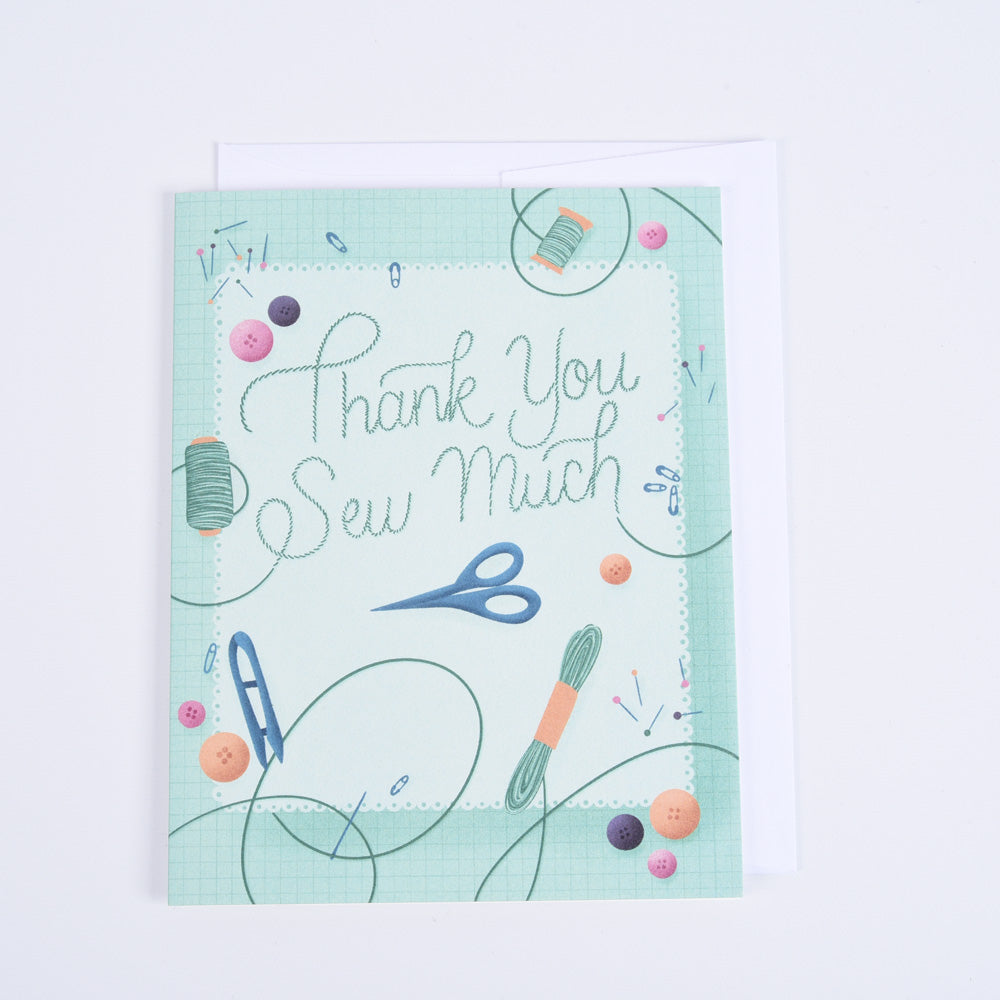 A folded greeting card with a colorful design featuring sewing-related images such as scissors, thread, and fabric, with the text &