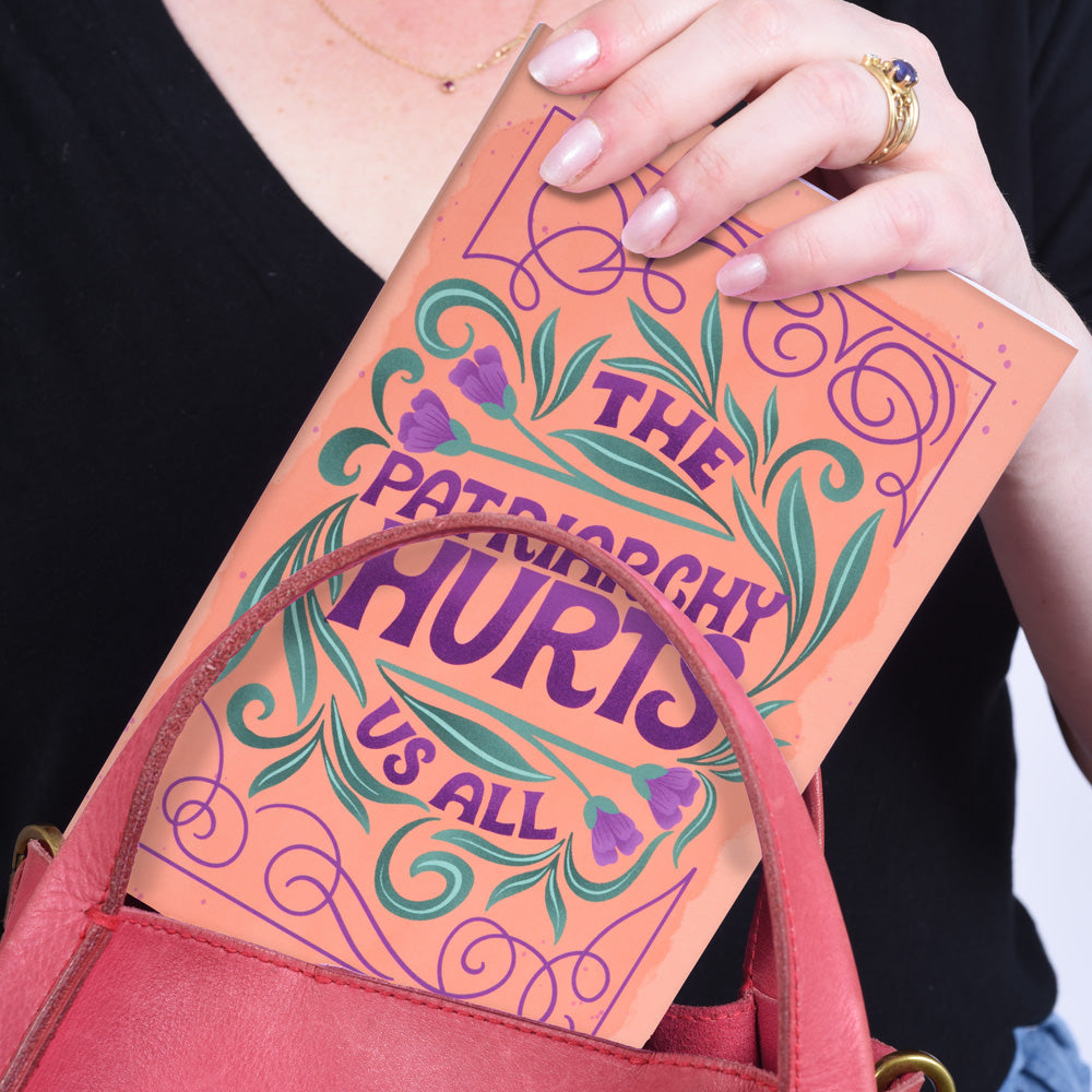 hand putting an orange journal with text "the patriarchy hurts us all" into a pink purse