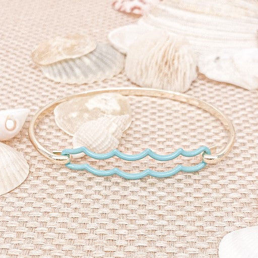 the beach and back double aqua wave tension bangle in gold tone on neutral color woven textured table top with white seashells scattered in the background