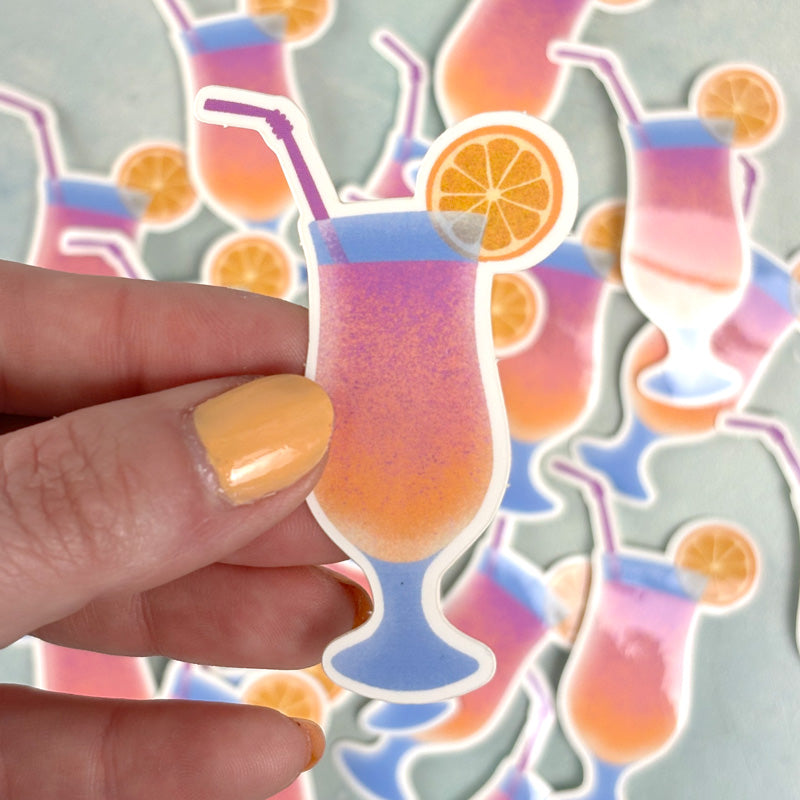 hand holding A sticker depicting a colorful cocktail with a swirl of orange liquid, accompanied by a straw and an orange slice