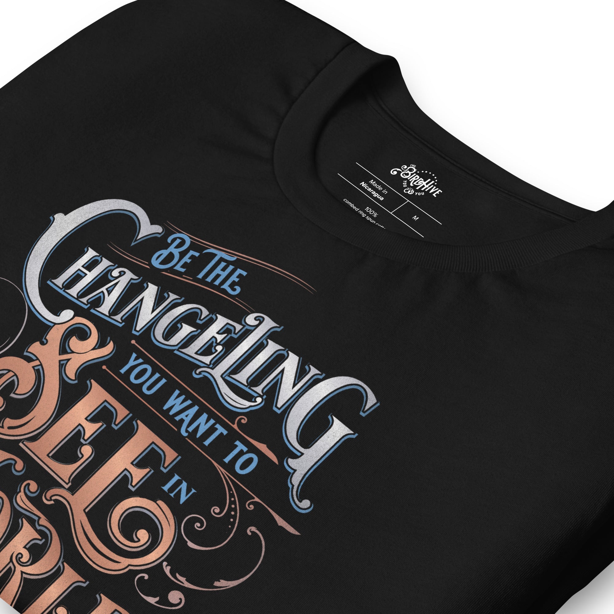 folded Black unisex fit tee with design “Be the Changeling you want to see in the world” in silver, bronze and blue lettering surrounded by intricate decals