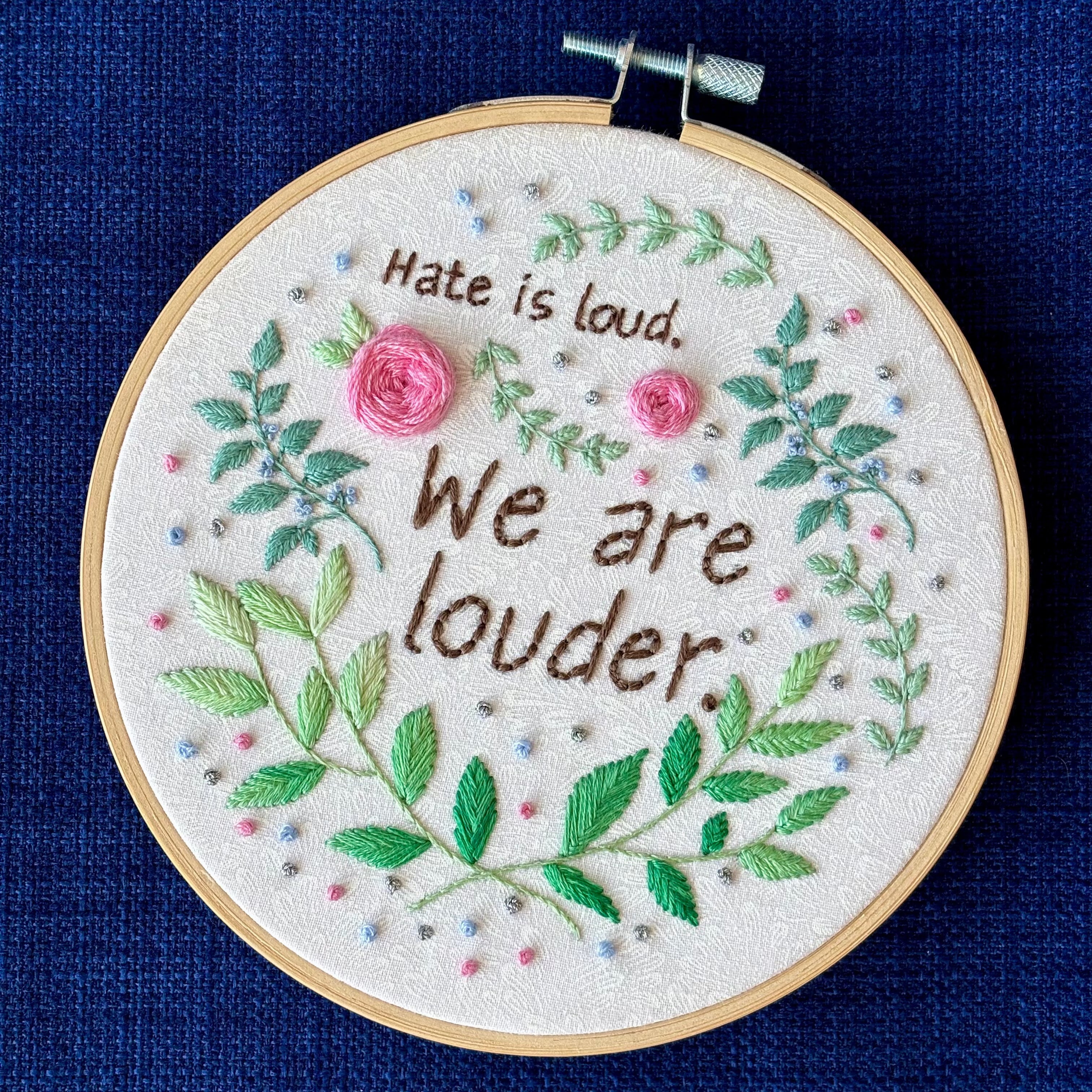 "Hate is loud. We are louder." Pride quote hand embroidered in chocolate brown. Surrounded by green leaves & vines, with pink roses, blue berries, & metallic silver dots. Sewn on white-on-white cotton. Framed in light wood embroidery hoop as wall art.