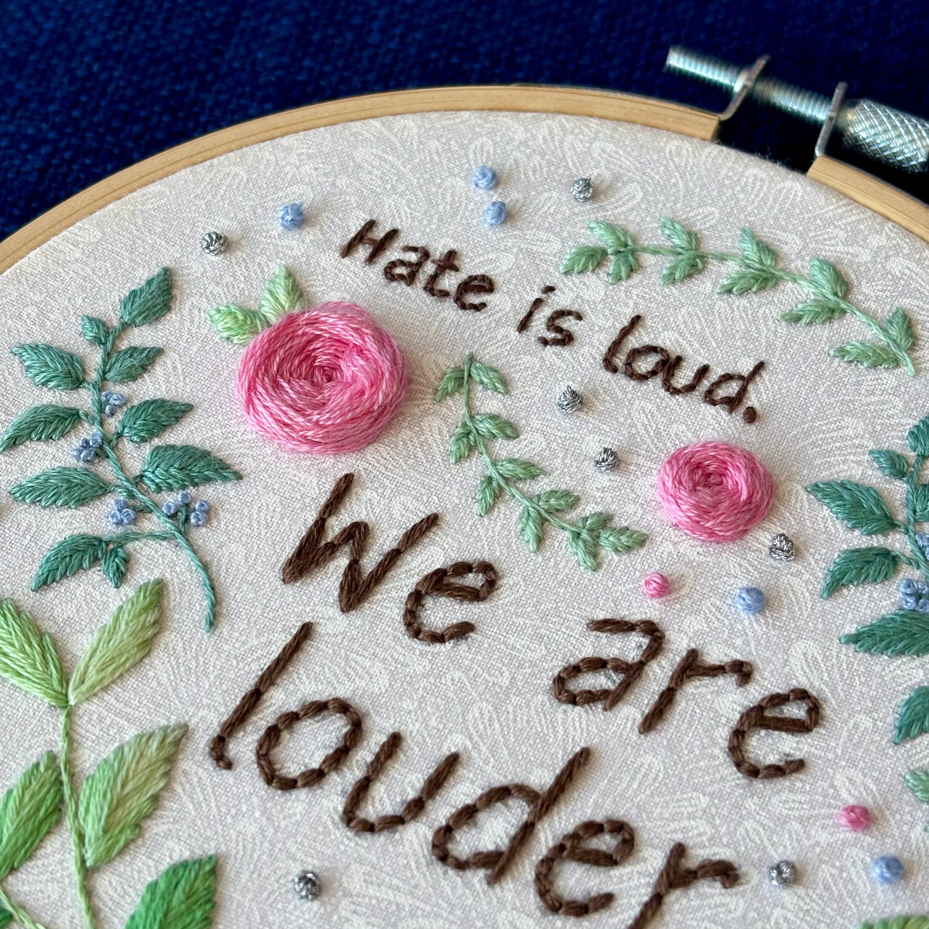 "Hate is loud. We are louder." Pride quote hand embroidered in chocolate brown. Surrounded by green leaves & vines, with pink roses, blue berries, & metallic silver dots. Sewn on white-on-white cotton. Framed in light wood embroidery hoop as wall art.