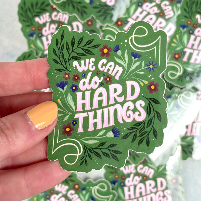 hand holding a floral sticker with lettering "we can do hard things" on a green background