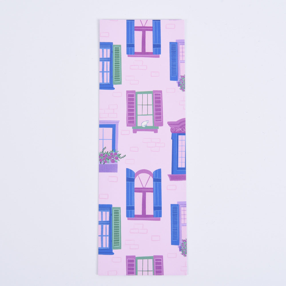 A bookmark featuring a pattern of whimsical city scenes with windows, flower boxes, and a cat on a pink brick backdrop.