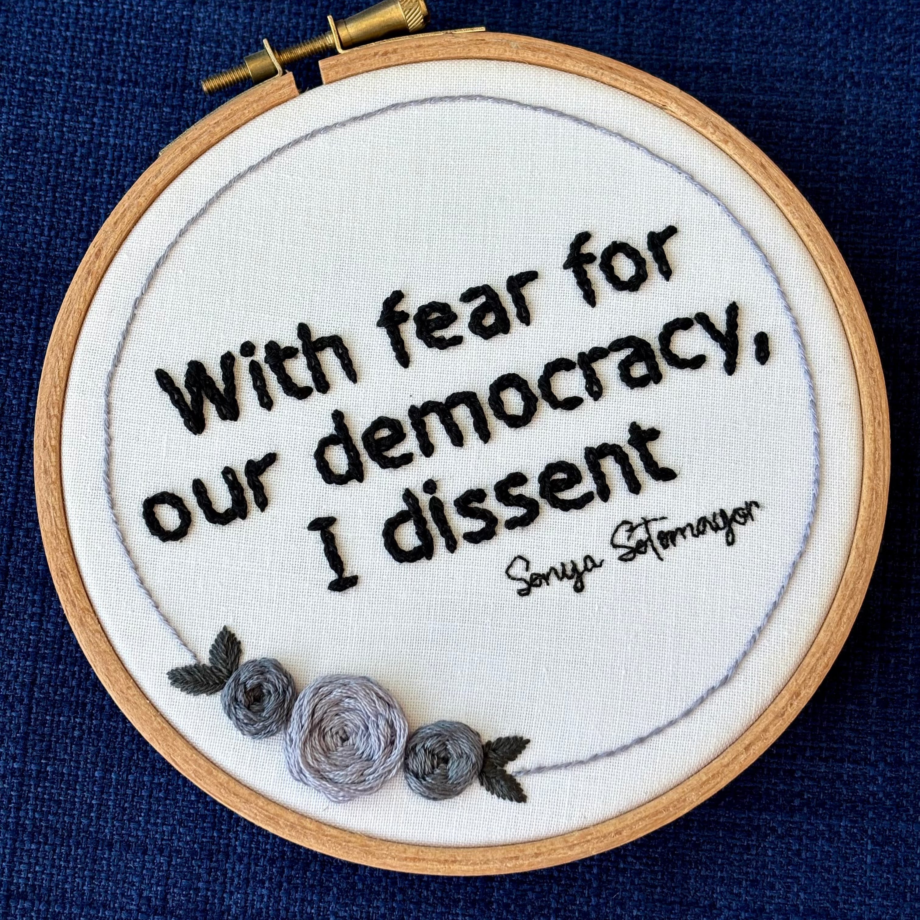 "With fear for our democracy, I dissent. Sonia Sotomayor" Quote hand embroidered in a strong sans-serif font in black embroidery thread. A floral wreath of light and medium gray roses surrounds the text. Fabric is white cotton. Embroidery is displayed as wall art in a wood embroidery hoop.