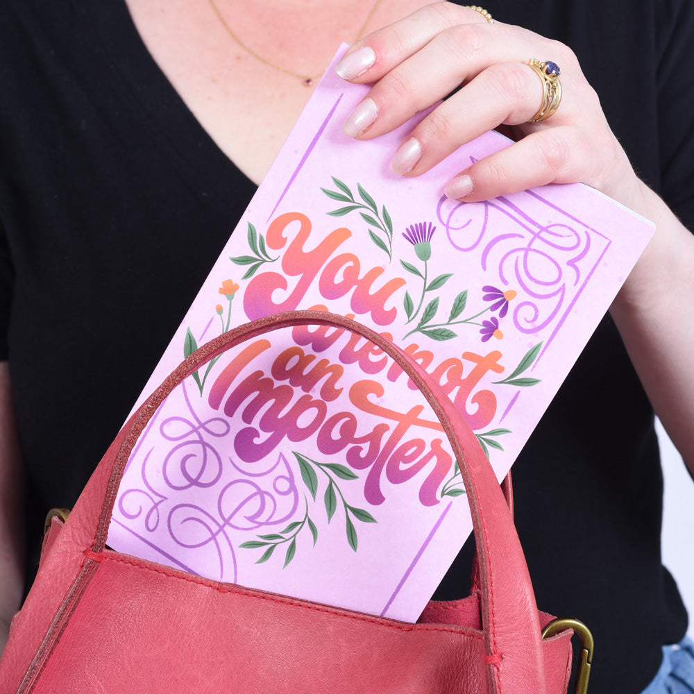 hand putting a pink notebook with flowers and text "you are not an imposter" into a pink purse