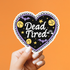 A hand with white manicured nails holds a heart-shaped sticker against a solid orange background. The sticker features a black background with the words &