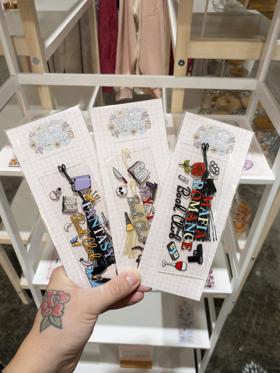 Hand holding three acrylic bookmarks with designs and text, against a blurred indoor background.