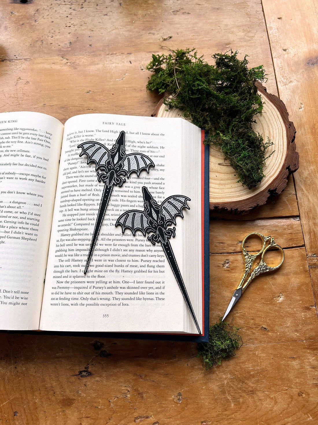 Dragon-shaped bookmarks on an open book with a pair of scissors and a piece of moss on a wooden surface.