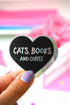 A hand with lavender and green glitter nail polish holds a heart-shaped black sticker that reads &