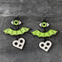 A pair of gothic-style bat dangle earrings with bright green wings, a green eye with a black heart, and a white skull heart charm, displayed on a dark gray textured background.