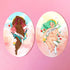 Two oval art prints or stickers displayed side-by-side on a solid pink background. The left print features a dark-skinned woman with pink hair and the text &