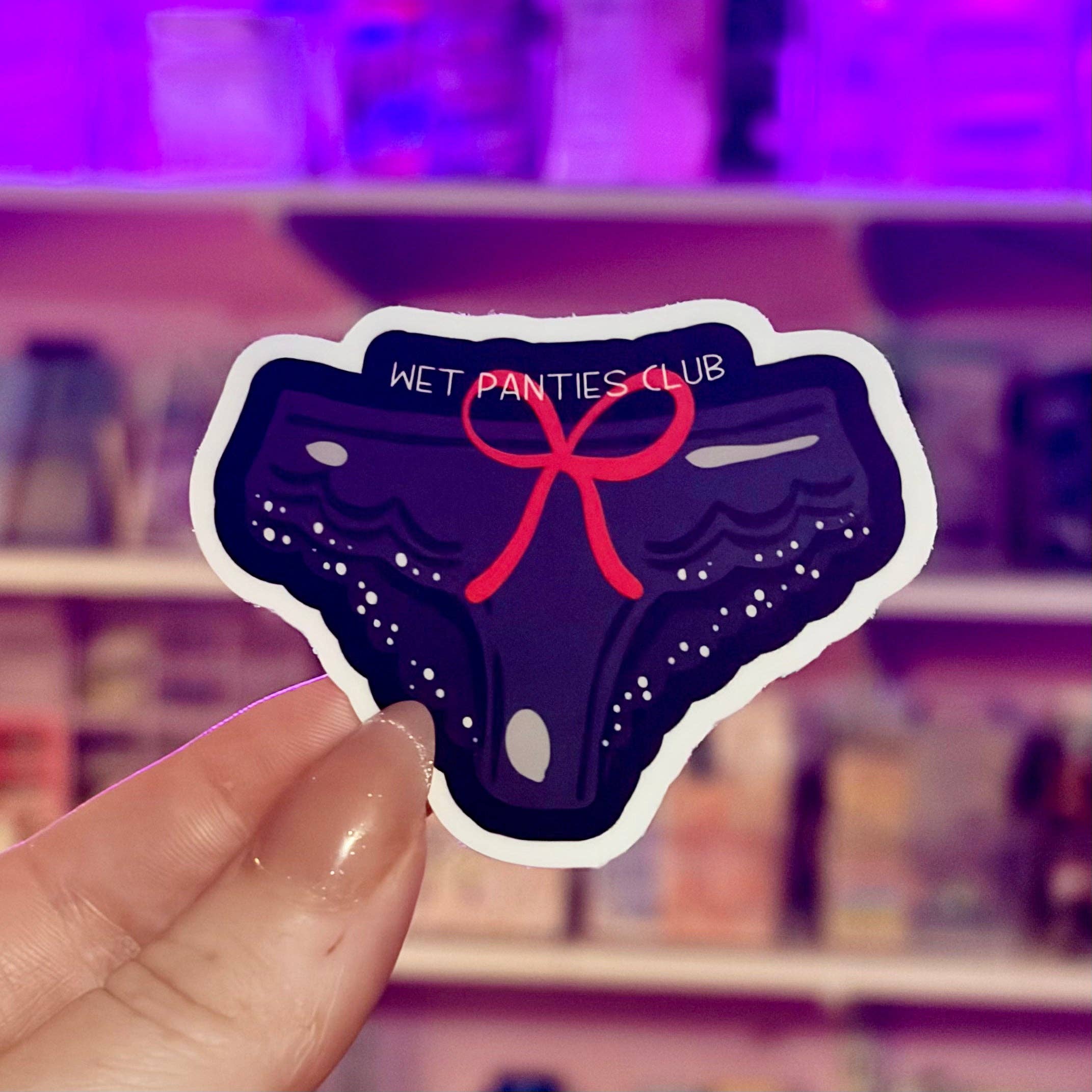 Sticker of a pair of wet panties with a red bow held in front of a blurred store shelf.