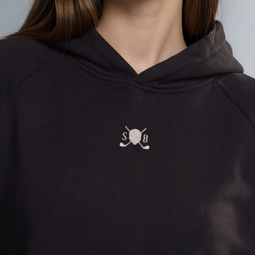 A woman modeling a black pullover hoodie with a small white embroidered cat logo on the chest, paired with dark gray jeans, against a neutral gray studio background.