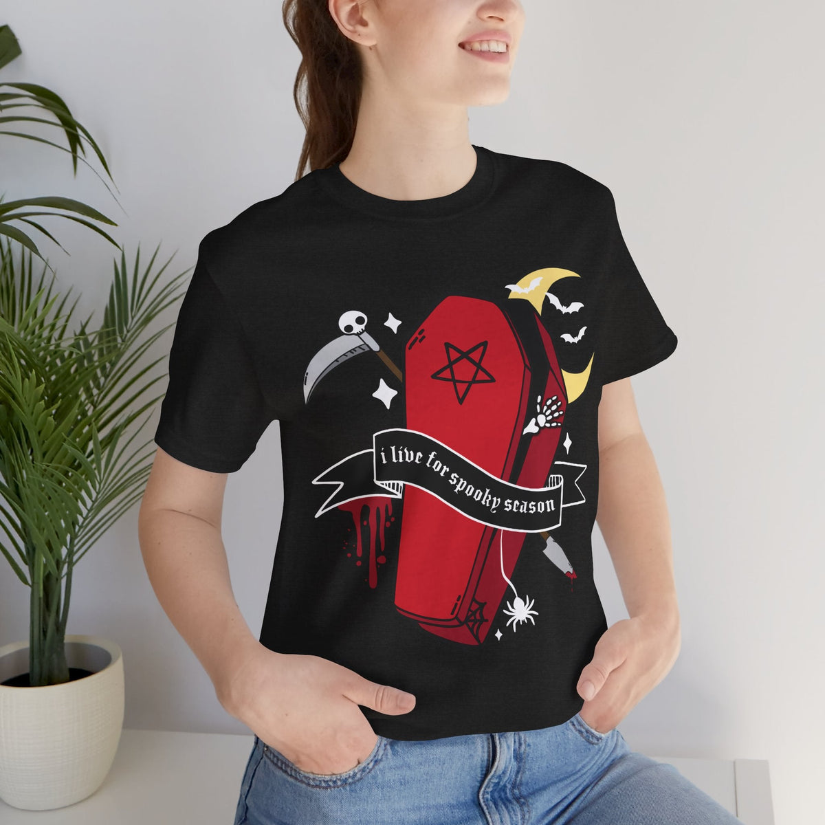 A woman wearing a black short-sleeve graphic t-shirt with a gothic Halloween design featuring a red coffin, scythe, spider, and banner that reads &
