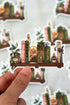Sticker of a shelf with books, plants, and candles held by a hand against a blurred background.