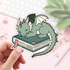 Sticker of a green dragon on a book held by a hand with a blurred background