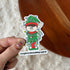 A hand holds a die-cut sticker featuring a cartoon elf wrapped in Christmas lights, with the text &