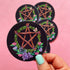 A hand with mint green nail polish holds a round black sticker featuring a wooden pentagram inside a green leaf wreath with pink and purple flowers. Three more identical stickers are stacked in the background against a solid pink backdrop.