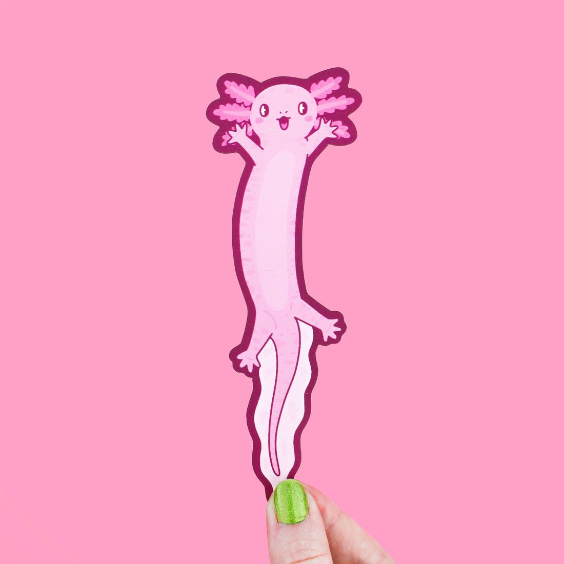Pink axolotl bookmark held by a hand with green nail polish against a pink background