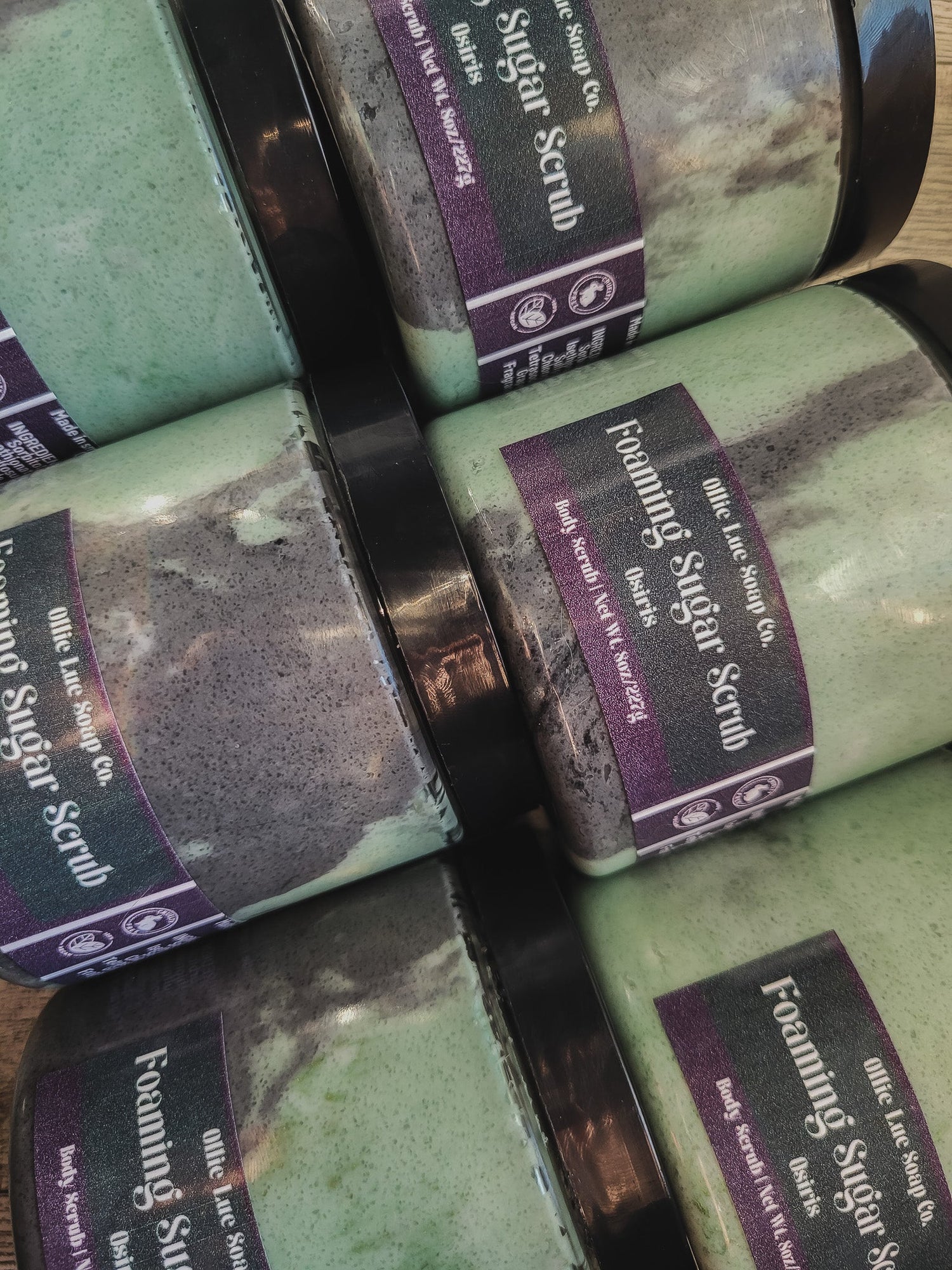 Green and purple Foaming Sugar Scrub containers stacked on a wooden surface.