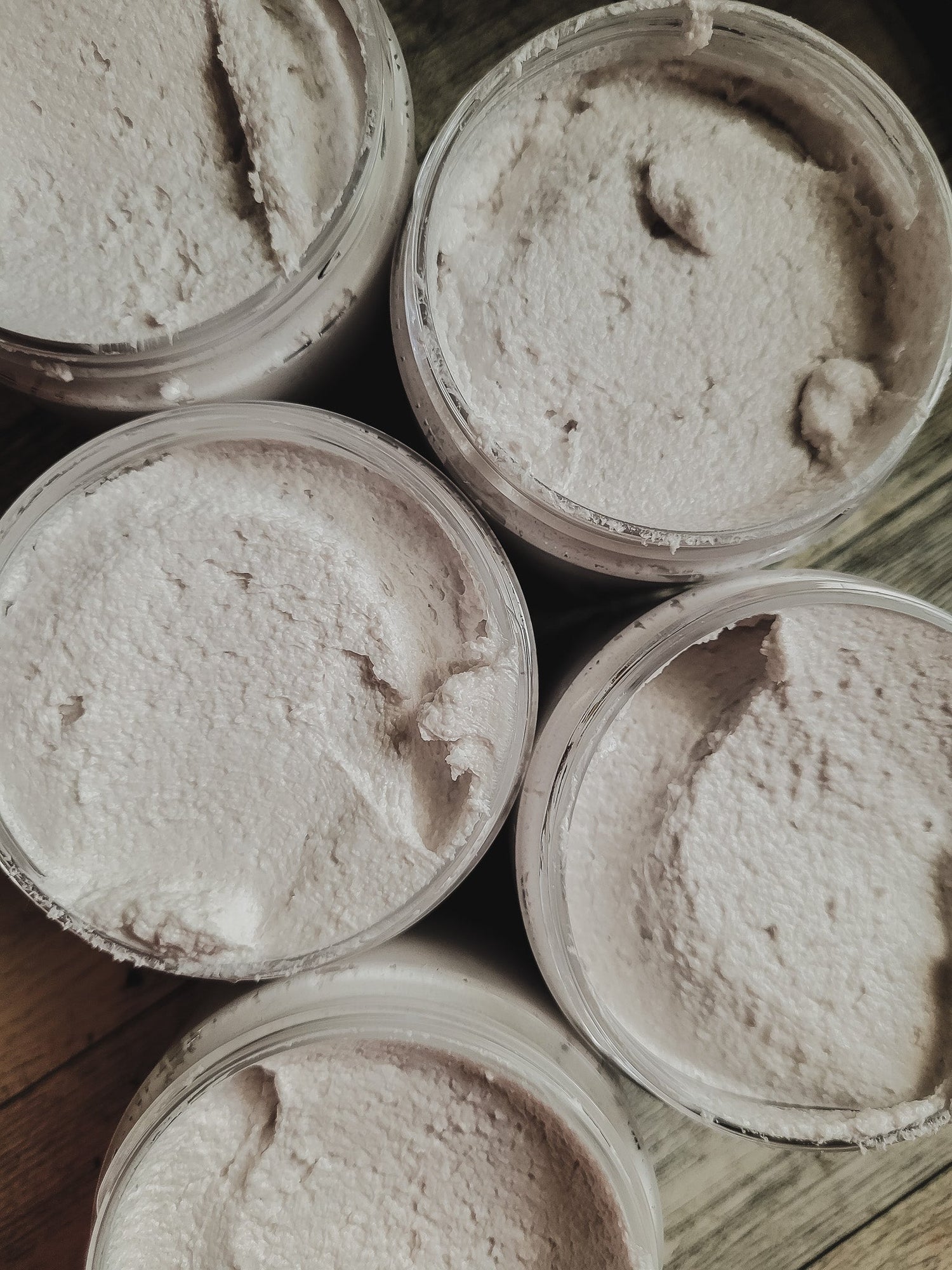 Five containers of a light purple scrub on a wooden surface.