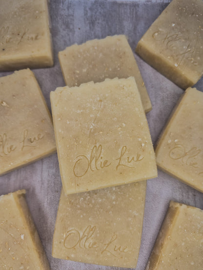 Several rectangular bars of natural soap in a warm beige-tan color, arranged on a textured gray surface. The soap bars are embossed with the brand name &