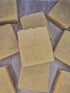 Several rectangular bars of natural soap in a warm beige-tan color, arranged on a textured gray surface. The soap bars are embossed with the brand name &