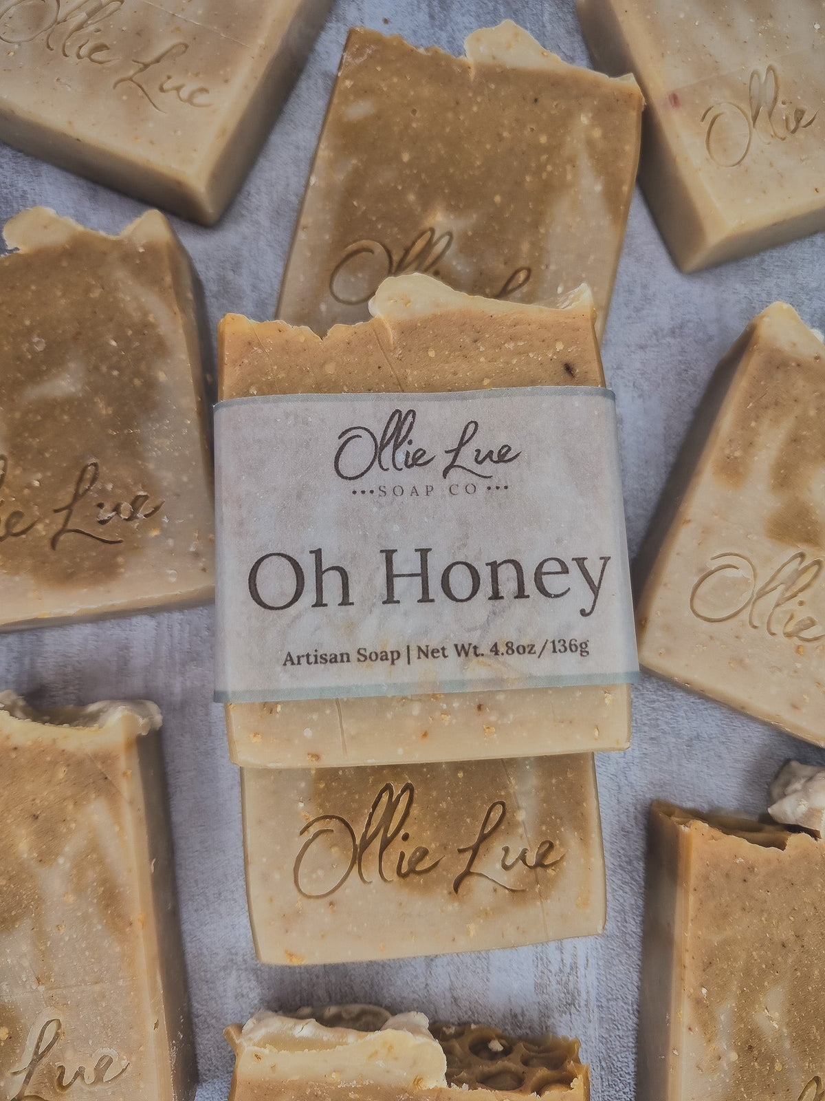 A stack of handmade artisan soap bars in a warm honey beige color, with a central bar wrapped in a paper label that reads &