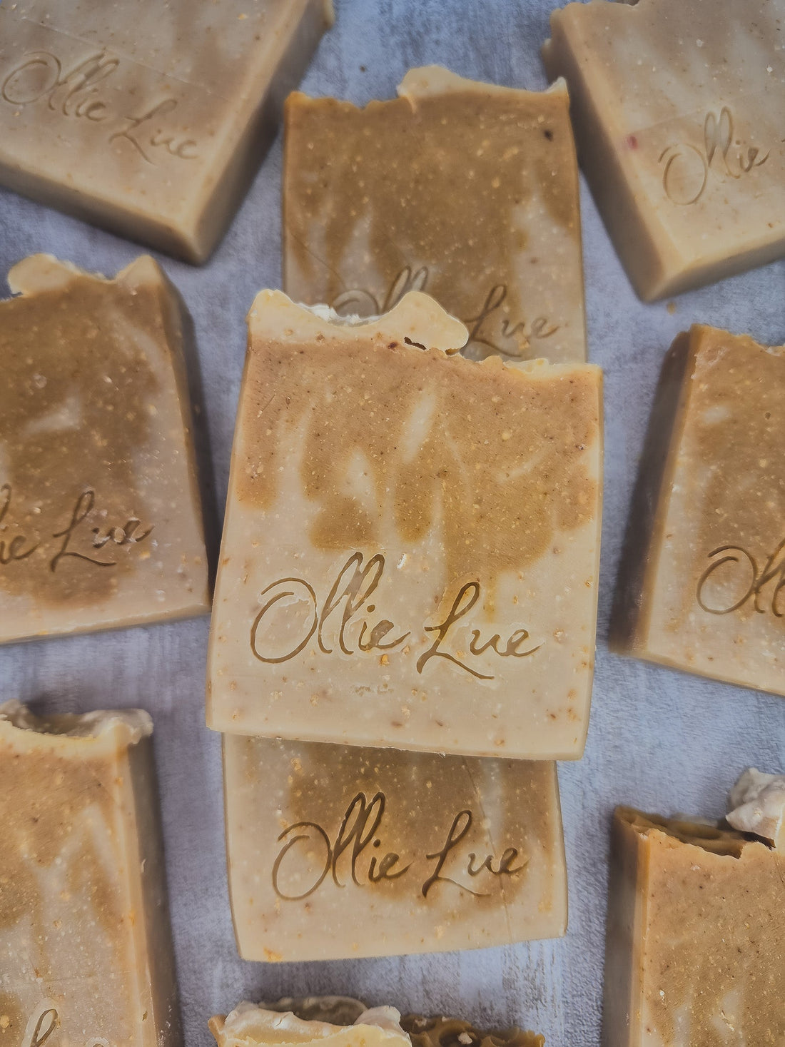 Several handmade rectangular bars of soap in warm tan and beige tones are arranged on a light gray surface. The center bar is stacked on another, and the soap pieces are embossed with the brand name &