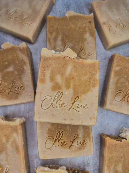 Several handmade rectangular bars of soap in warm tan and beige tones are arranged on a light gray surface. The center bar is stacked on another, and the soap pieces are embossed with the brand name &
