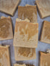 Several handmade rectangular bars of soap in warm tan and beige tones are arranged on a light gray surface. The center bar is stacked on another, and the soap pieces are embossed with the brand name &