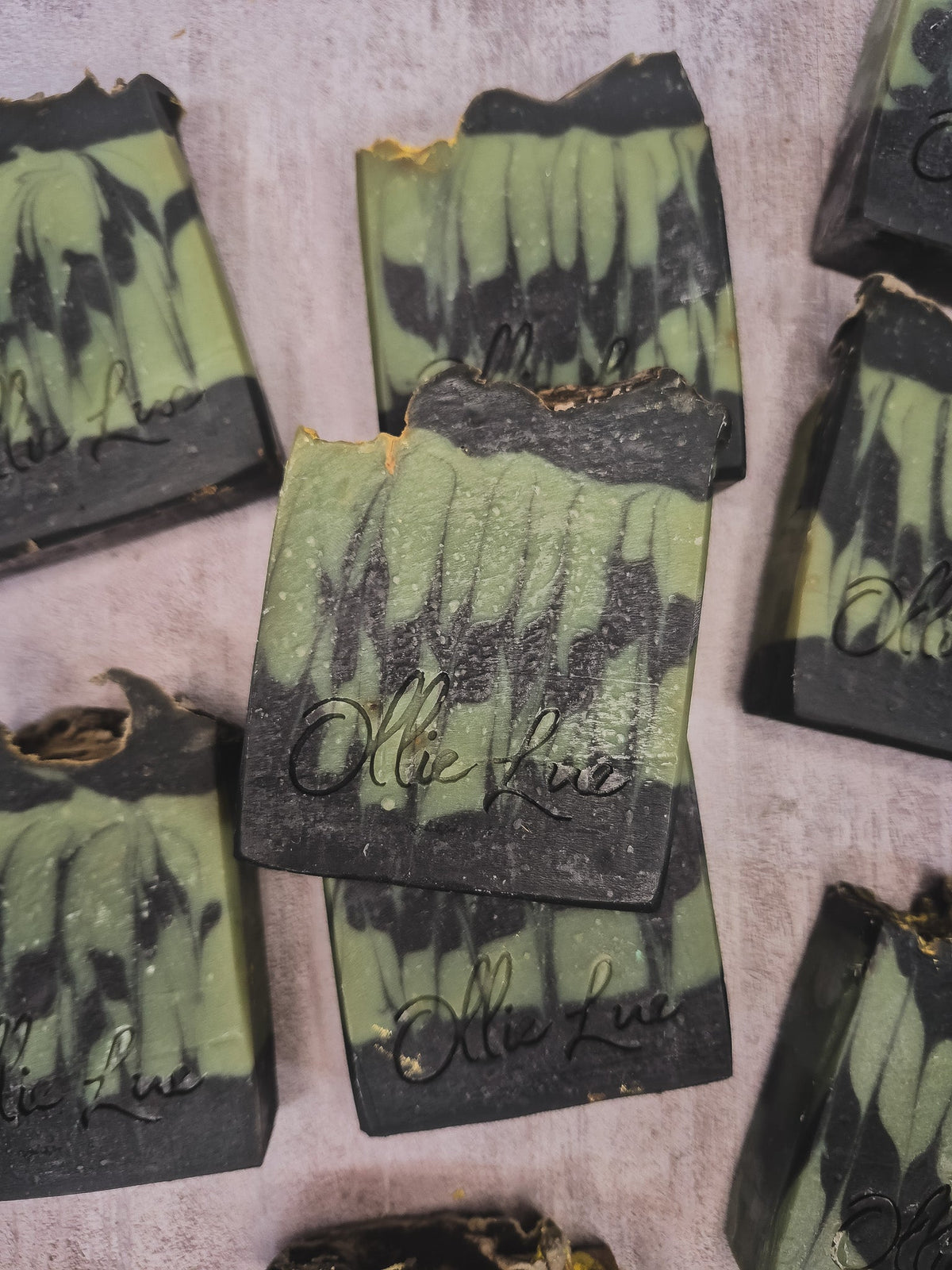 Several handmade soap bars with a green and black marbled pattern are arranged on a textured light gray surface. The center soap bar is upright and features the brand name &