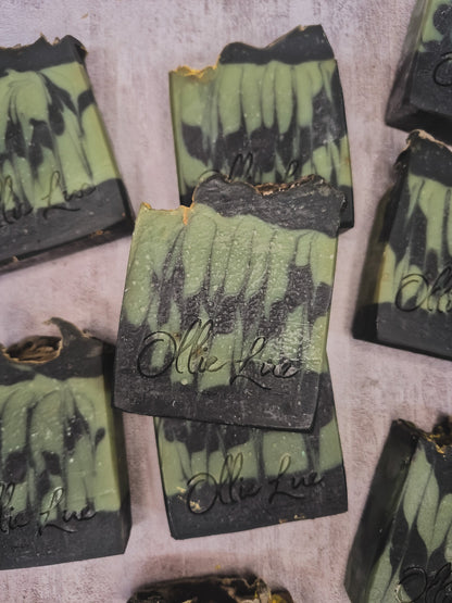 Several handmade soap bars with a green and black marbled pattern are arranged on a textured light gray surface. The center soap bar is upright and features the brand name &