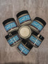 Six jars of Sweet Pits arranged in a circle on a textured gray surface, with one open jar in the center showing the creamy off-white oil.