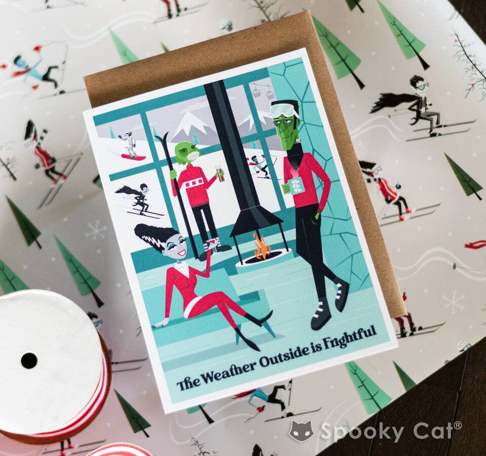 A square greeting card with a kraft envelope, featuring a whimsical winter illustration of three green-skinned characters in a snowy room. The card is placed on top of patterned wrapping paper with a spool of ribbon visible in the lower left.